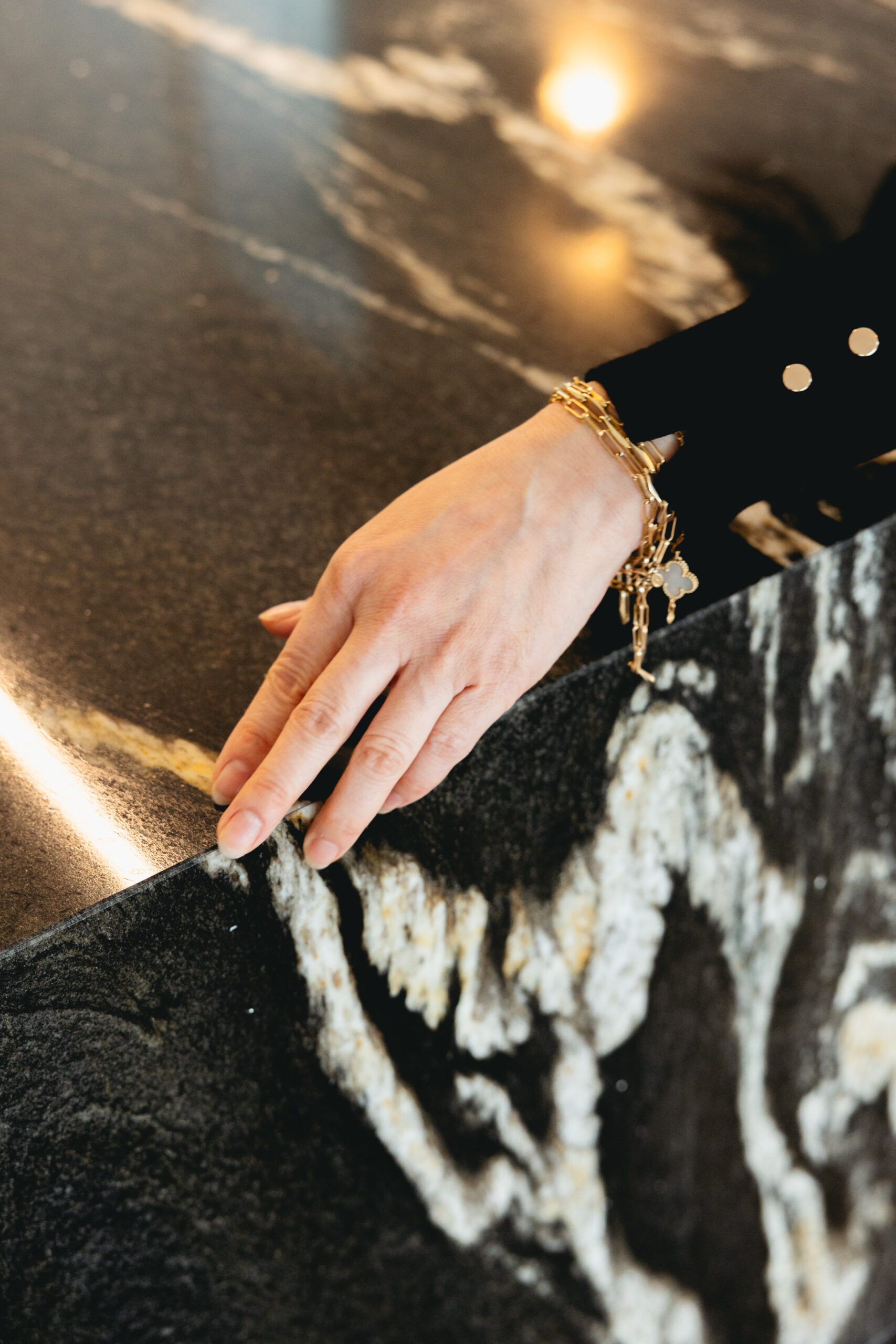 Hand resting on black marble countertop with gold veining, wearing stacked gold chain bracelets