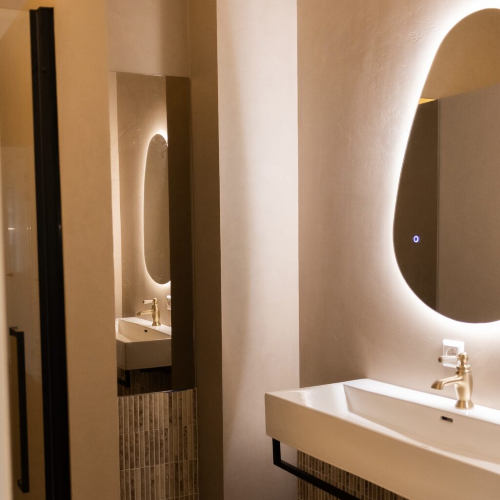 Luxury bathroom design with floating white sink, brass faucet, backlit oval mirror, and mosaic tile detail on warm plaster walls