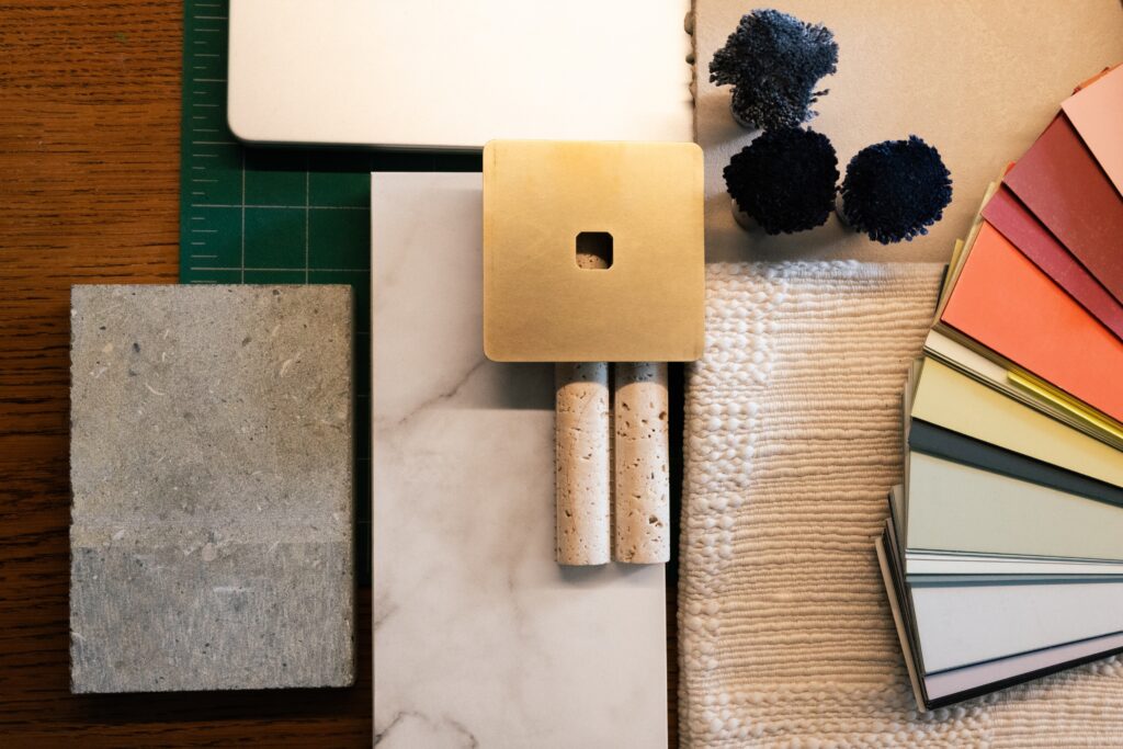 Interior design mood board with marble tile, concrete sample, brass cabinet hardware, woven fabric swatch, and color palette fan deck on a wooden desk