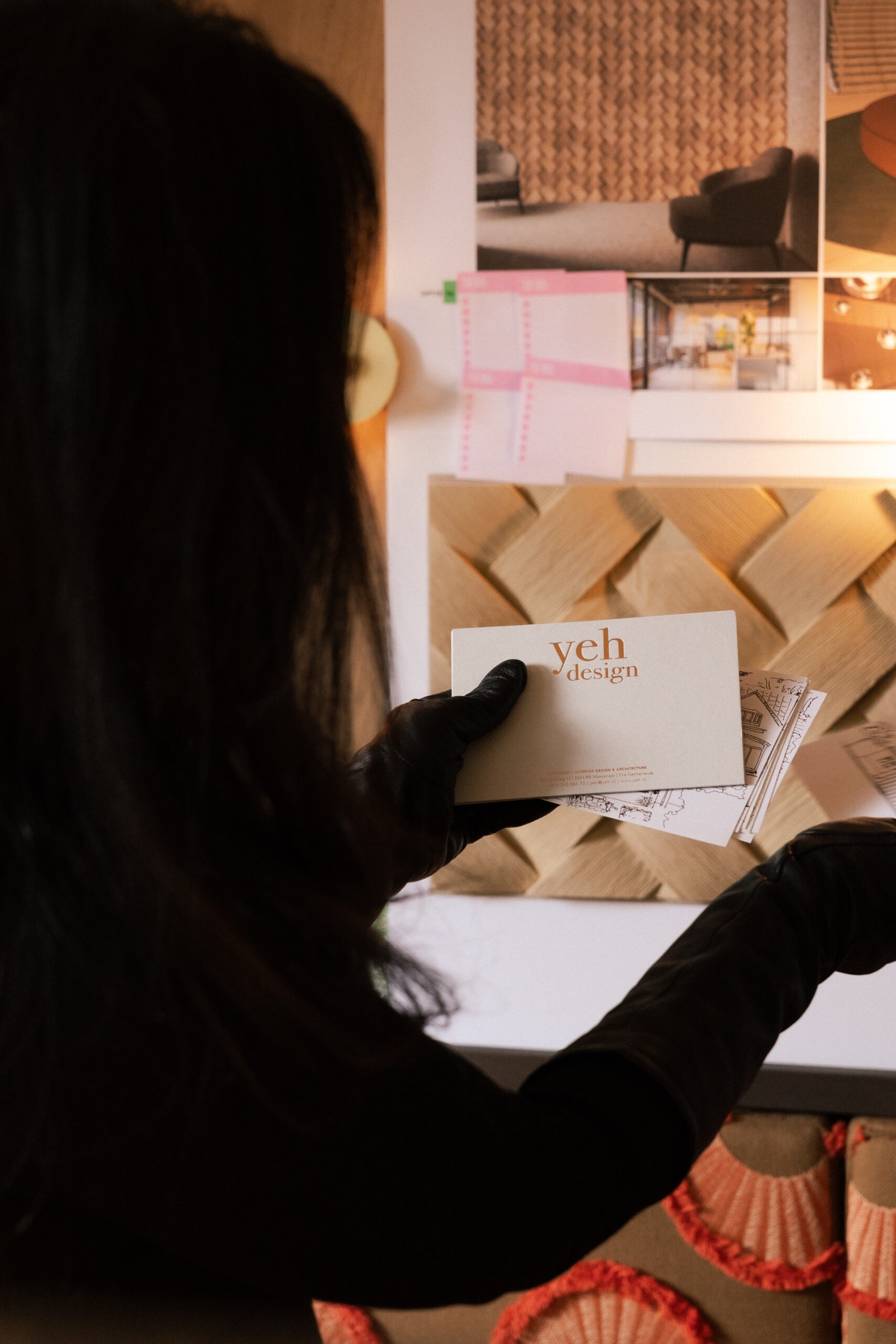 Interior designer holding Yeh Design business card in front of studio mood board with herringbone wood samples and interior design renders