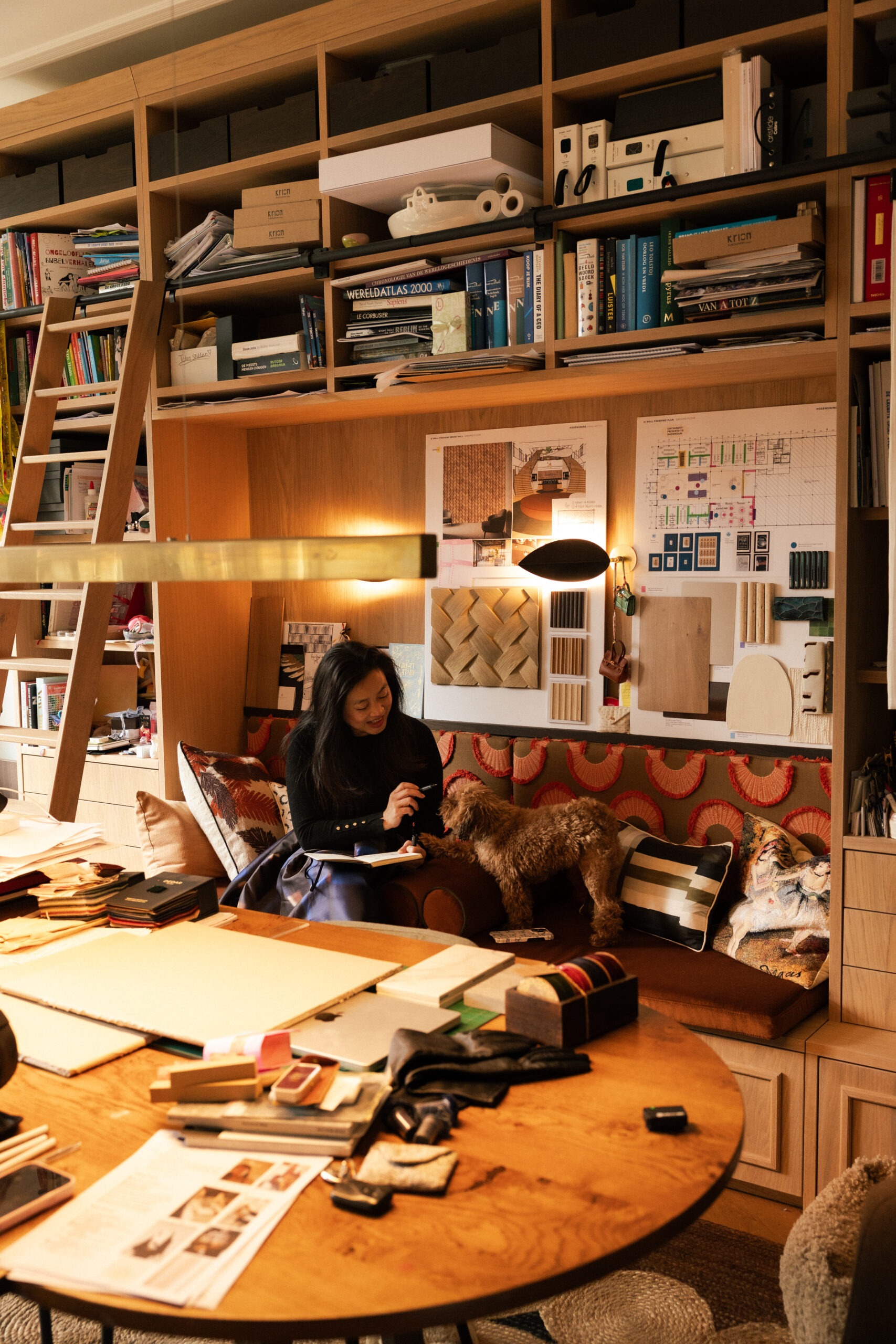 Interior designer writing in notebook on patterned sofa with dog beside her, material samples and floor plans pinned to studio wall behind her