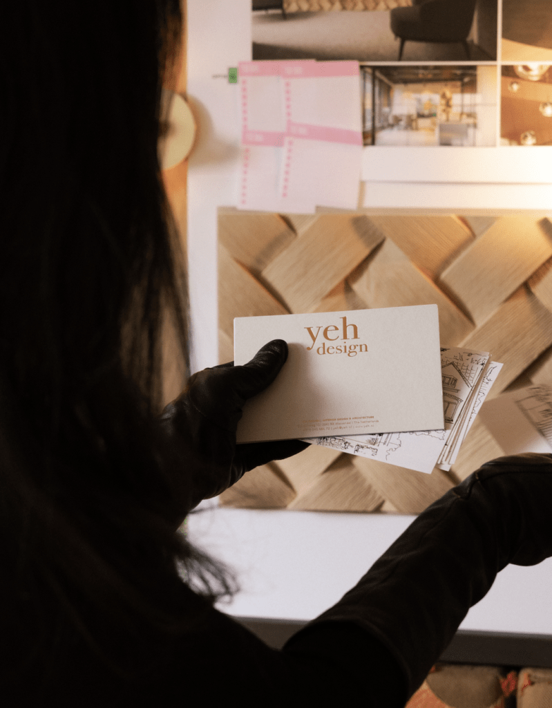 Interior designer holding Yeh Design business card in front of studio mood board with herringbone wood samples and interior design renders