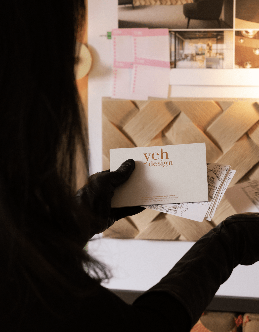 Interior designer holding Yeh Design business card in front of studio mood board with herringbone wood samples and interior design renders